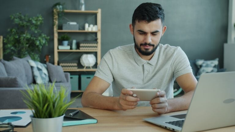 Man playing game on smartphone at desk with laptop