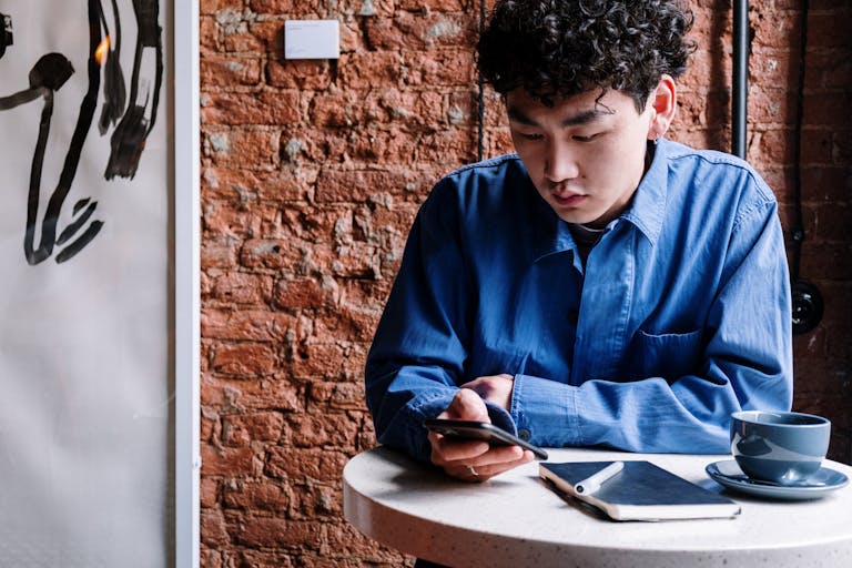 Asian man in blue shirt using cellphone at a café with a brick wall backdrop.