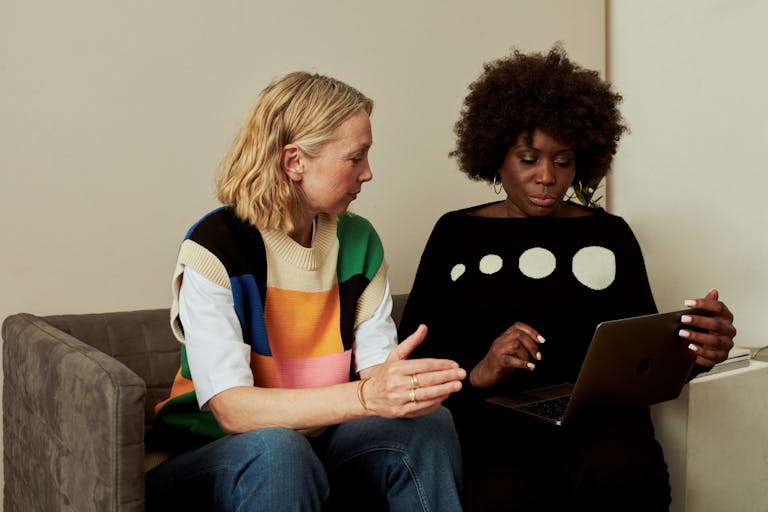 Two women in casual clothing discussing and using a laptop on a sofa, displaying teamwork and communication.